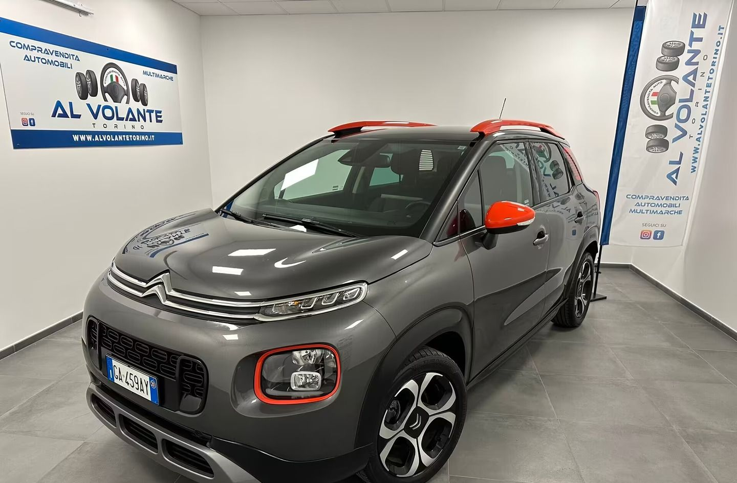 Una Citroën C1 compatta color argento parcheggiata in uno showroom luminoso con pareti bianche e striscioni del marchio sulle pareti laterali.