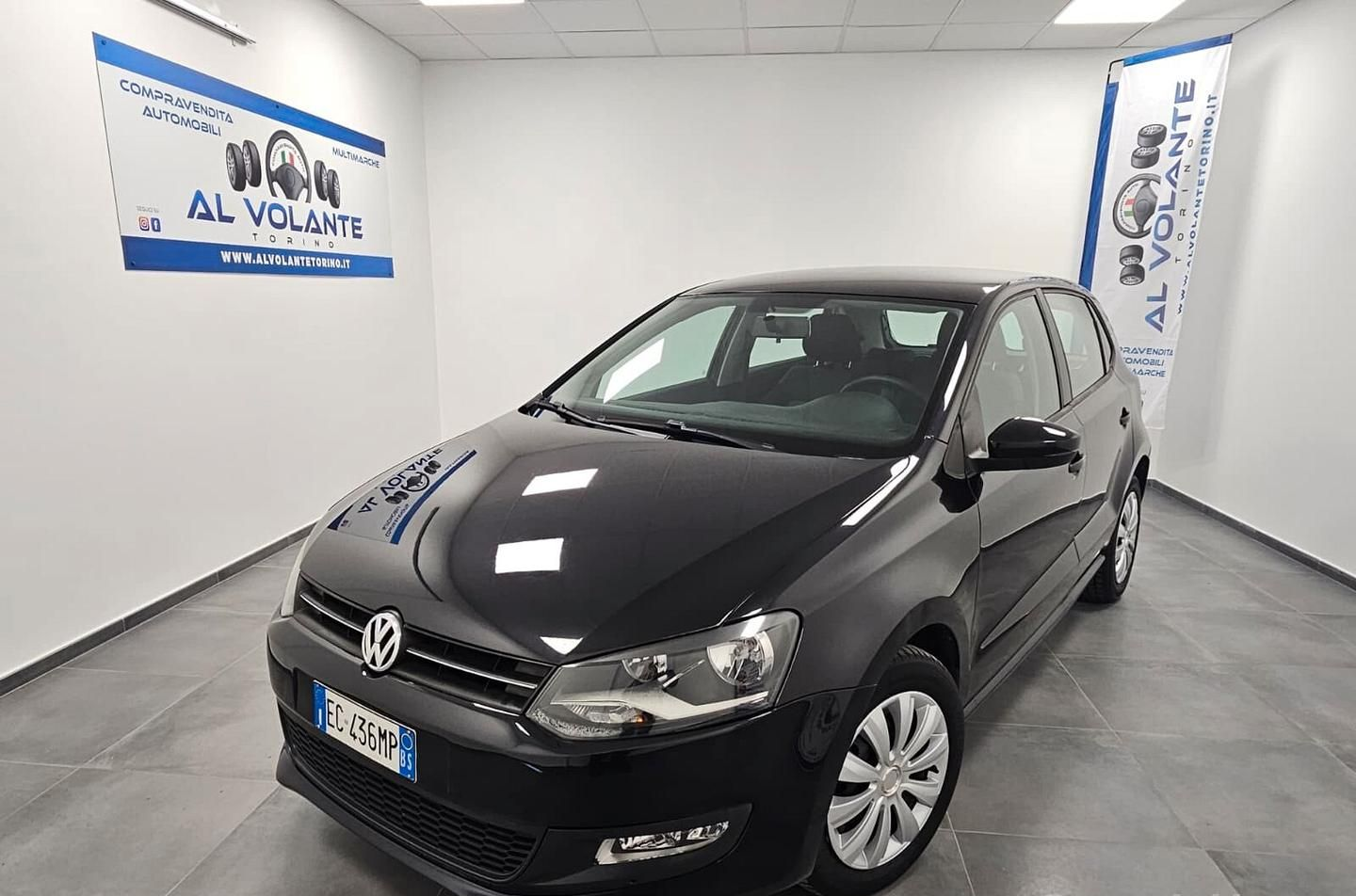 Una Peugeot 208 grigio scuro parcheggiata in uno showroom luminoso con pareti bianche e un'insegna con il logo sulla parete.