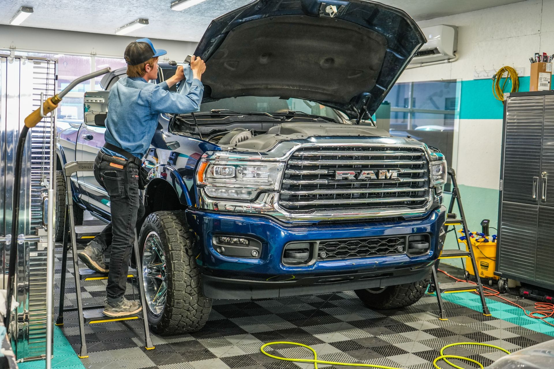 A man working on a blue RAM truck with its hood open inside a garage.