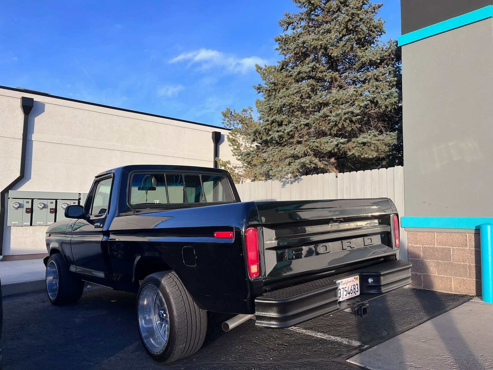 Black classic pickup truck parked near a building, silver wheels, blue sky.