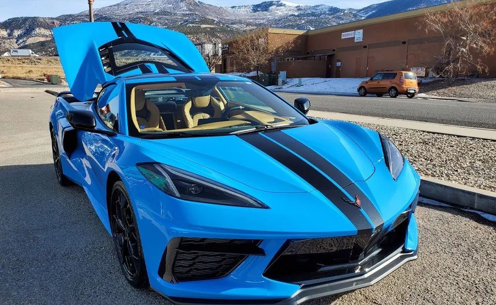 Blue Corvette sports car with black racing stripes and open hood; mountain backdrop.