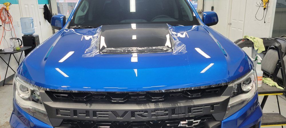 Blue Chevrolet pickup truck in a workshop, front view with hood wrapped in black and chrome grille visible