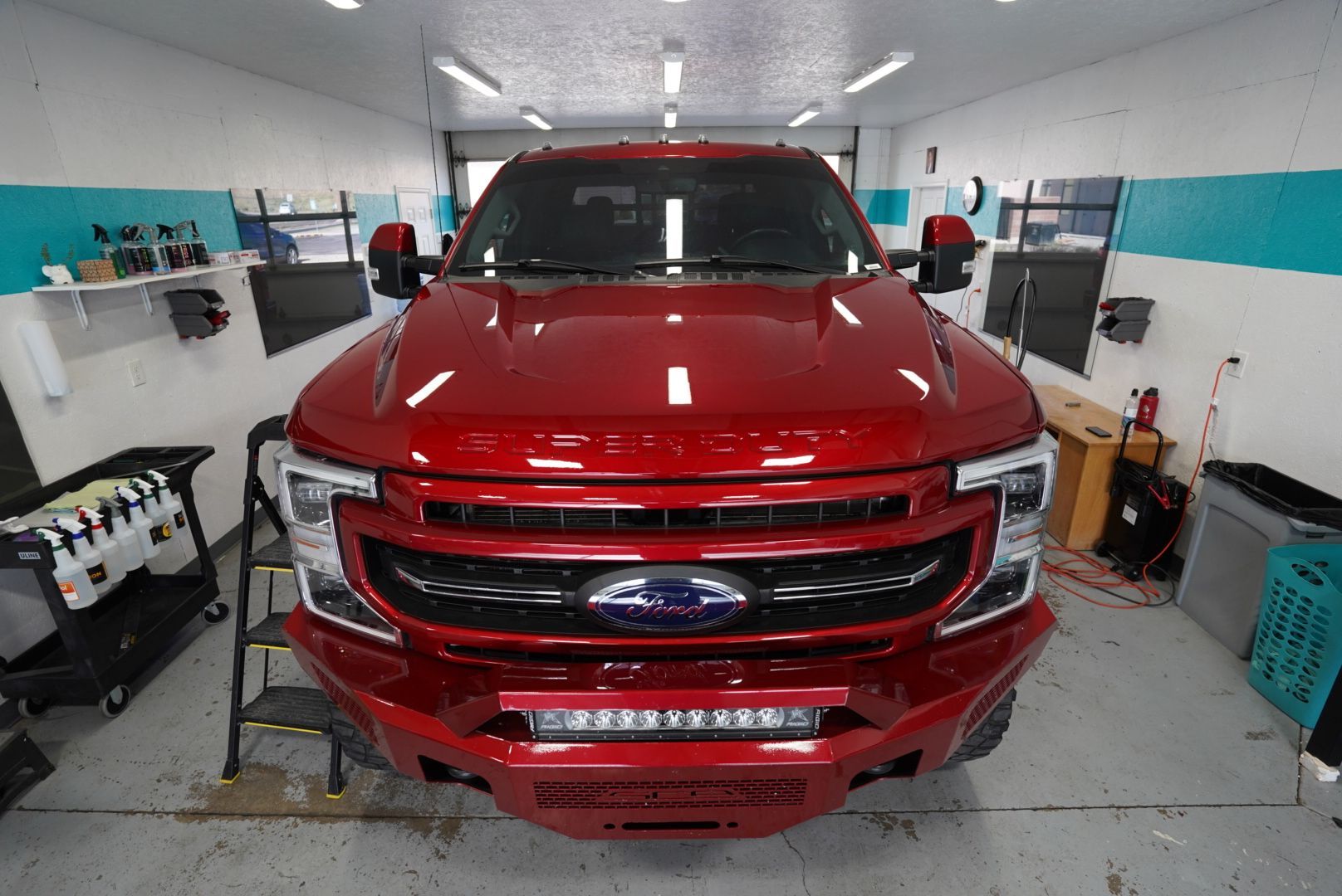 Red Ford truck in an auto shop, front bumper removed, surrounded by tools and equipment.