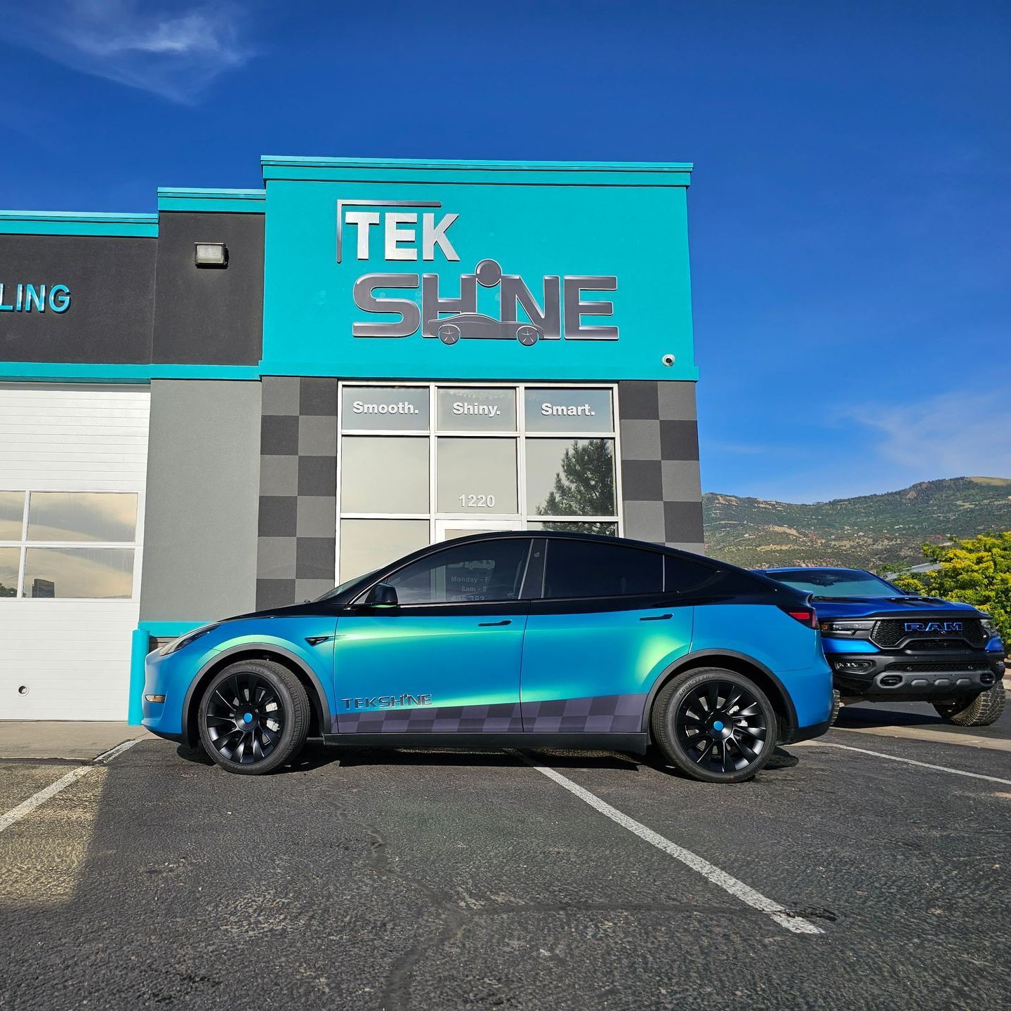 A teal Tesla parked in front of a turquoise auto detailing shop called Tek Shine. A truck is parked to its right.