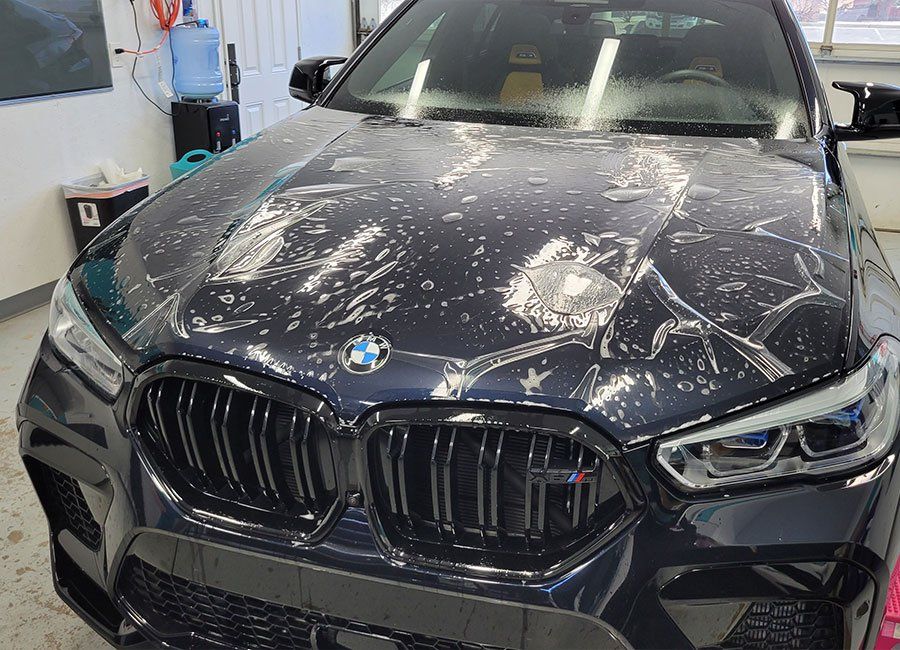 Black BMW sedan with heavy white splatter and scratches on the hood in a garage.