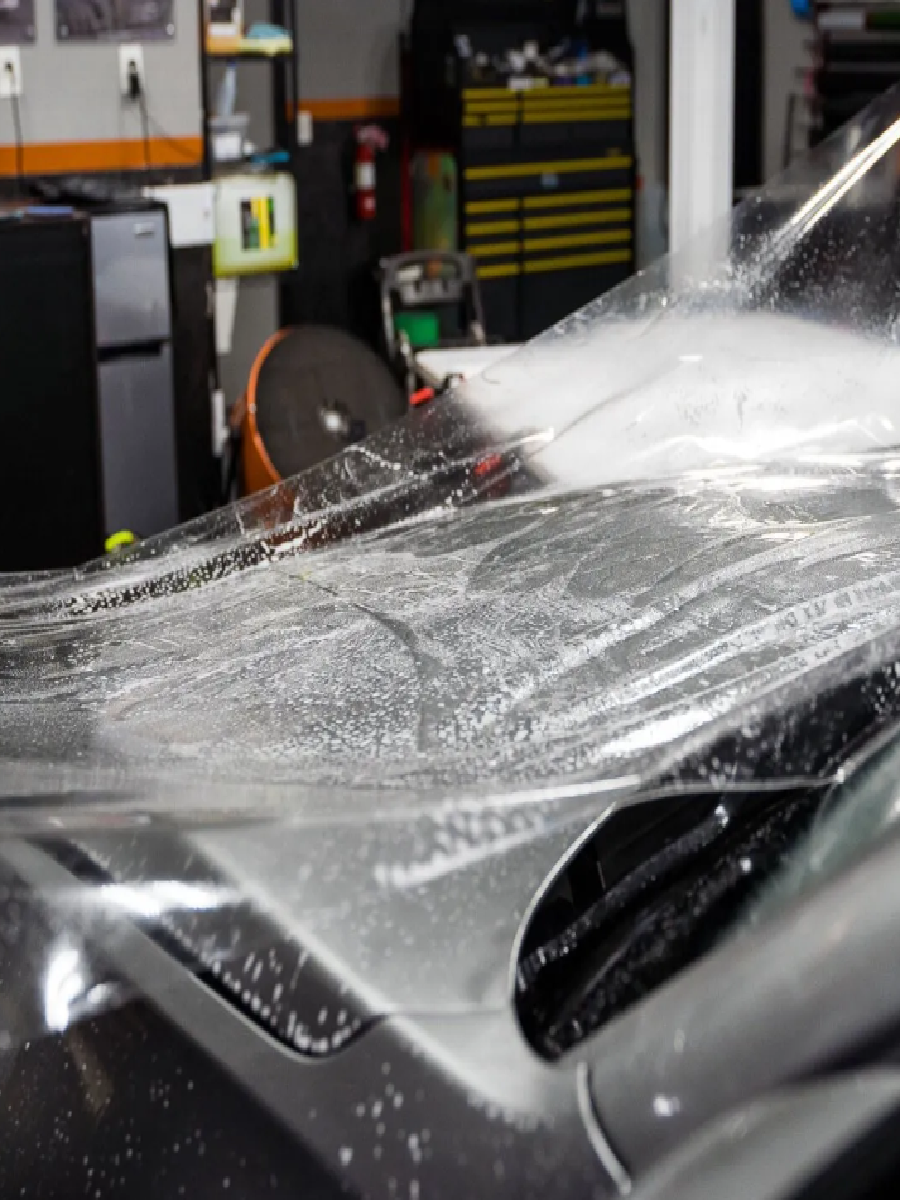A car hood being wrapped in clear plastic film in an auto shop, with tools and shelves in the background