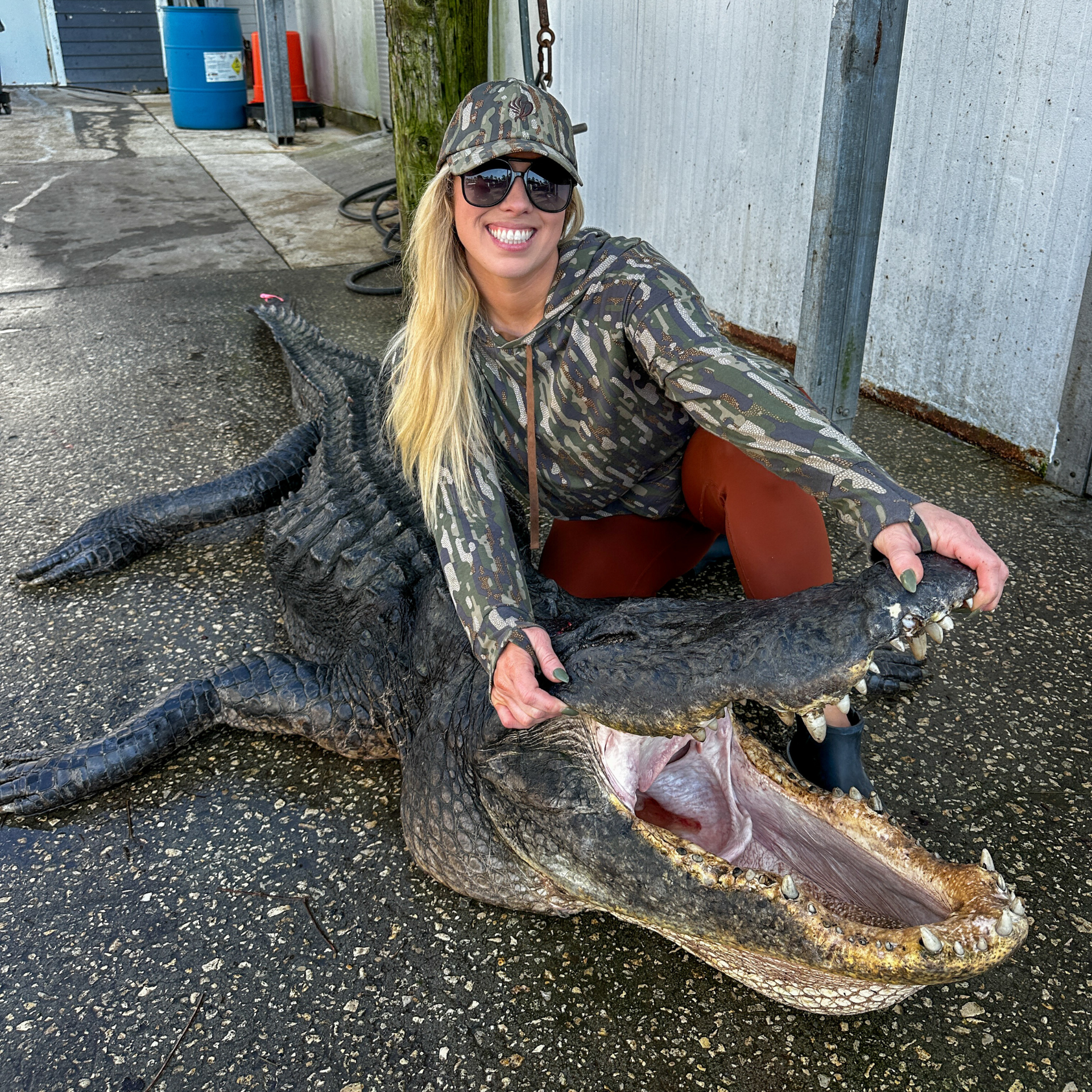 Woman in camo smiling, posing with a large alligator, its mouth open. Outdoors, concrete ground.