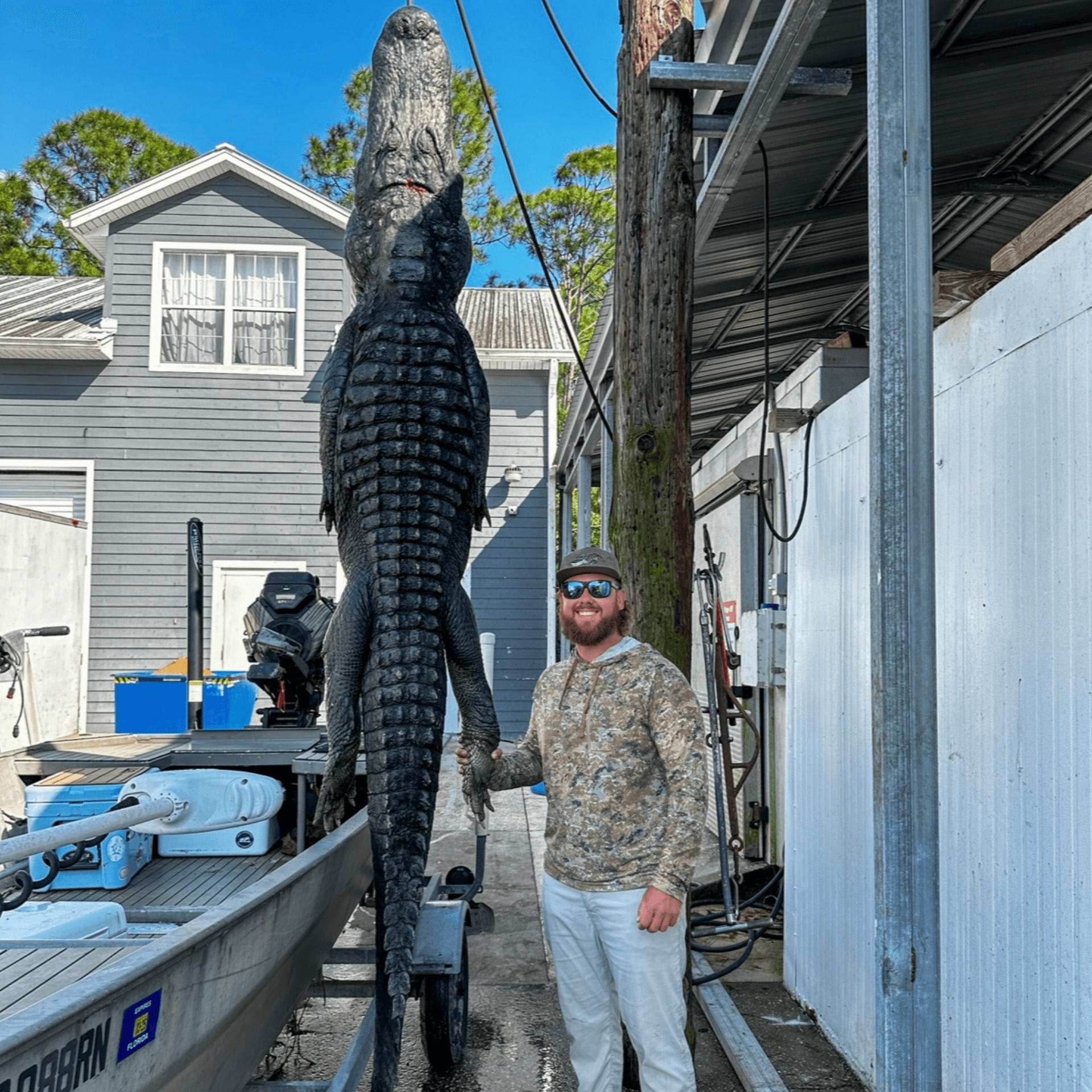 Man in camo stands next to a huge alligator suspended from a pole; outdoors, near a boat and a house.