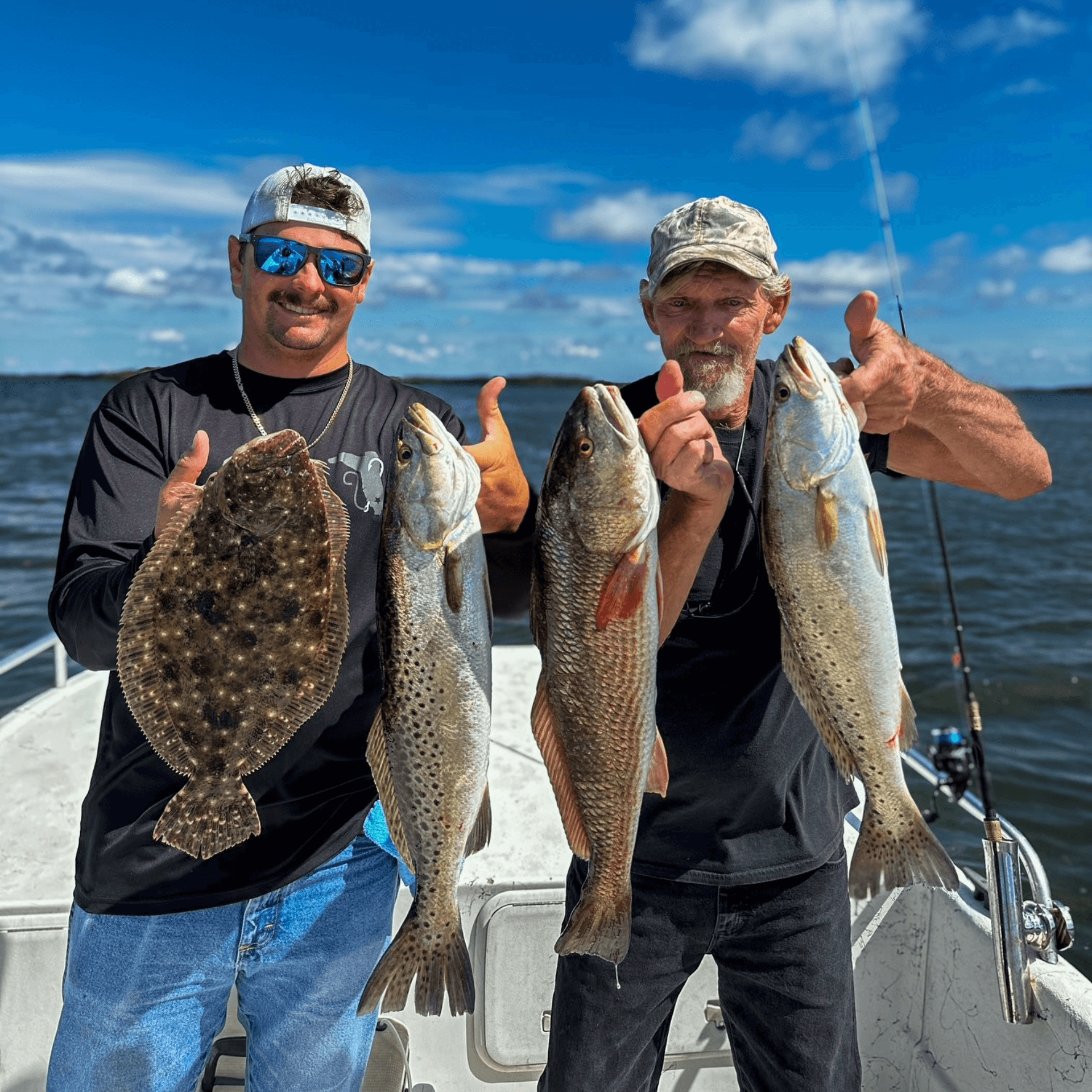 Two men are standing on a boat holding fish and giving a thumbs up.