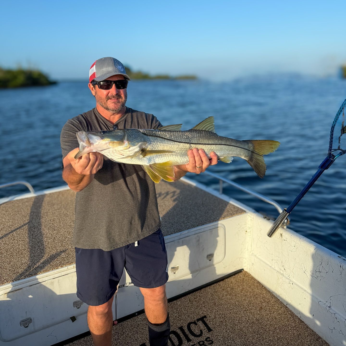 Man on boat holds a large snook fish. Blue water, gray sky, wearing hat and sunglasses.