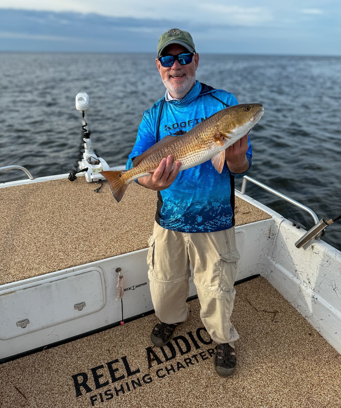 Man on boat holding up a redfish, blue shirt, khaki pants, ocean background.