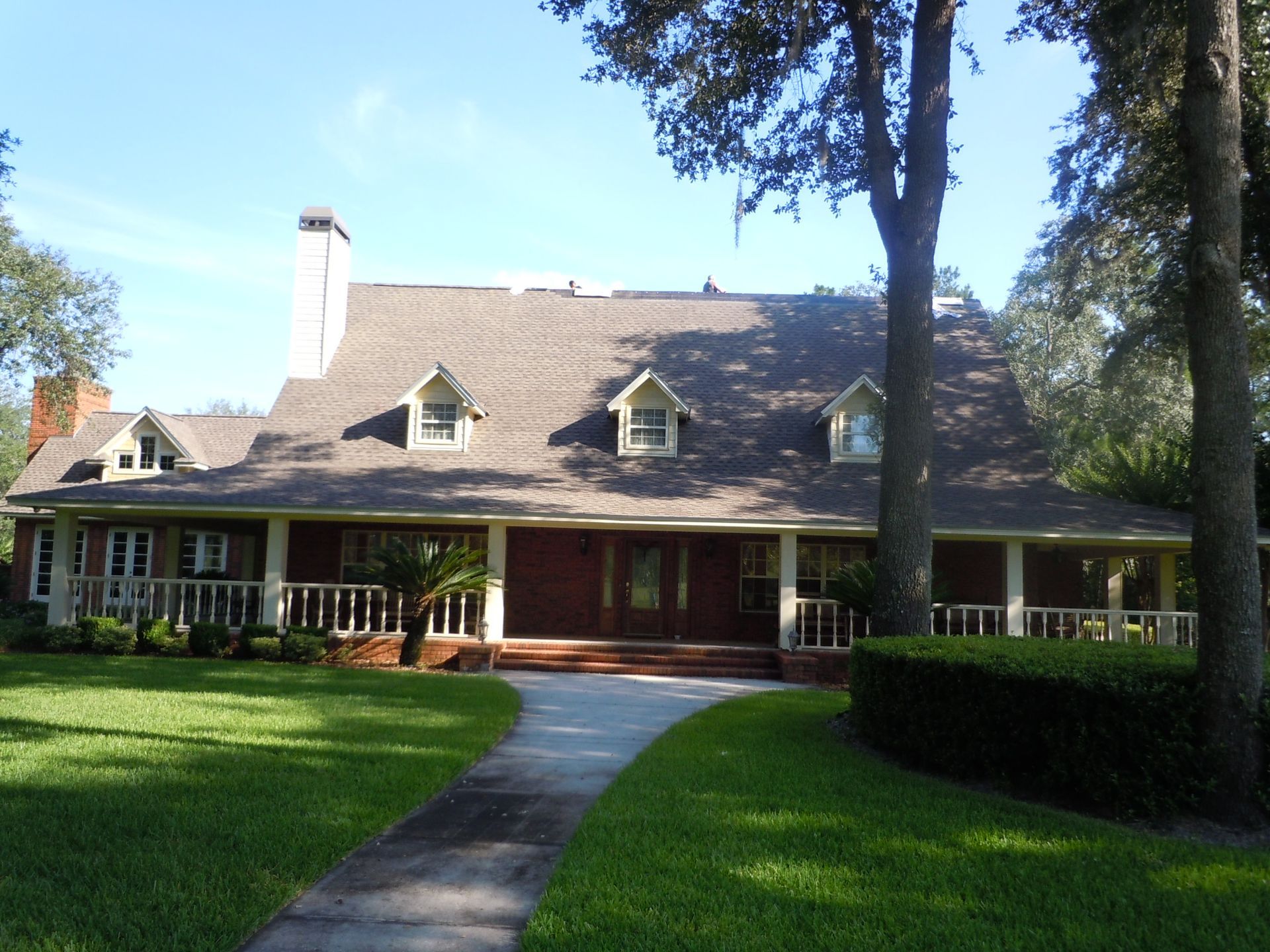 Large house with a wraparound porch, shingled roof, and front lawn under tall trees on a sunny day