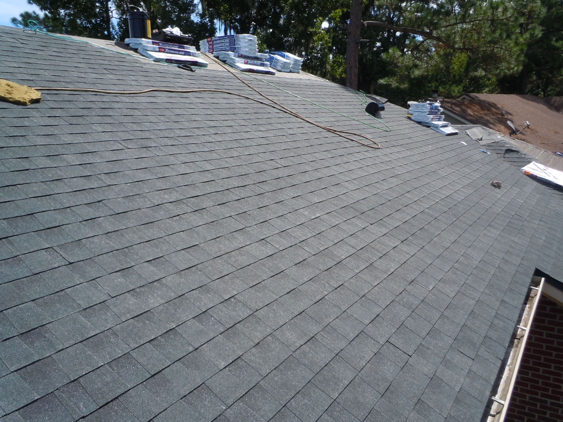 Gray shingled roof under repair with bundles of materials near the ridge and trees in the background