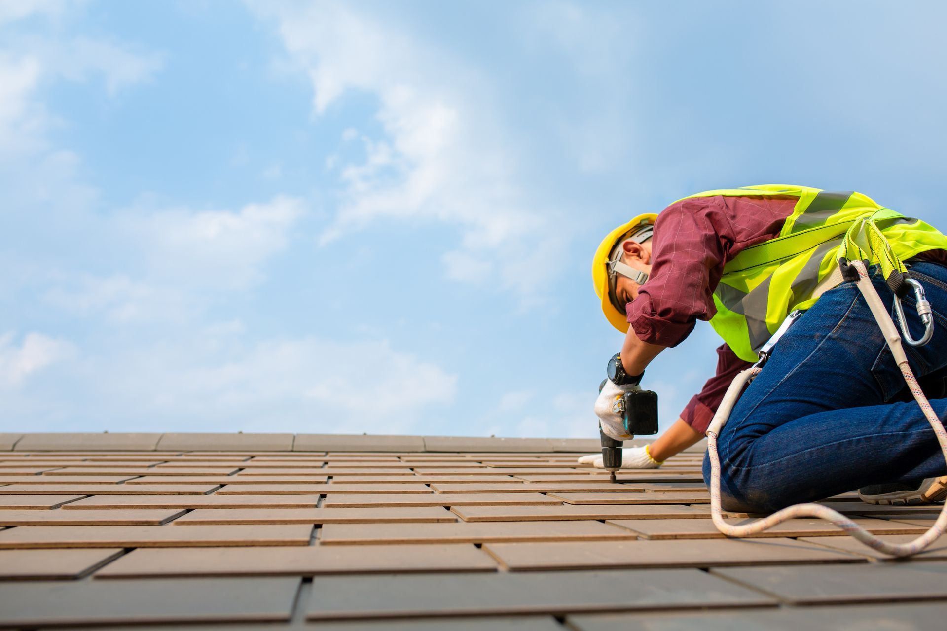 Worker in safety gear installing shingles on a roof under a blue sky