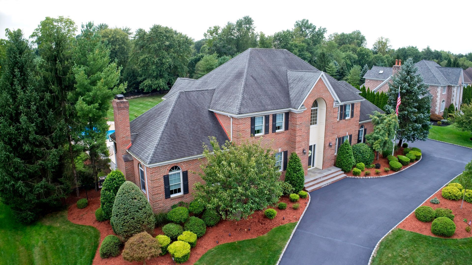 Brick house with gray roof, lush landscaping, and curved driveway in a wooded suburban setting