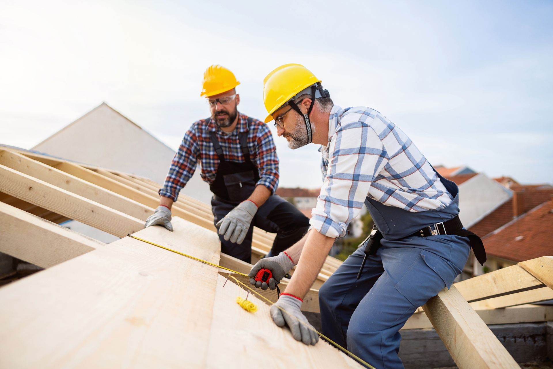 Two roofers in yellow hard hats measuring lumber on a rooftop construction site