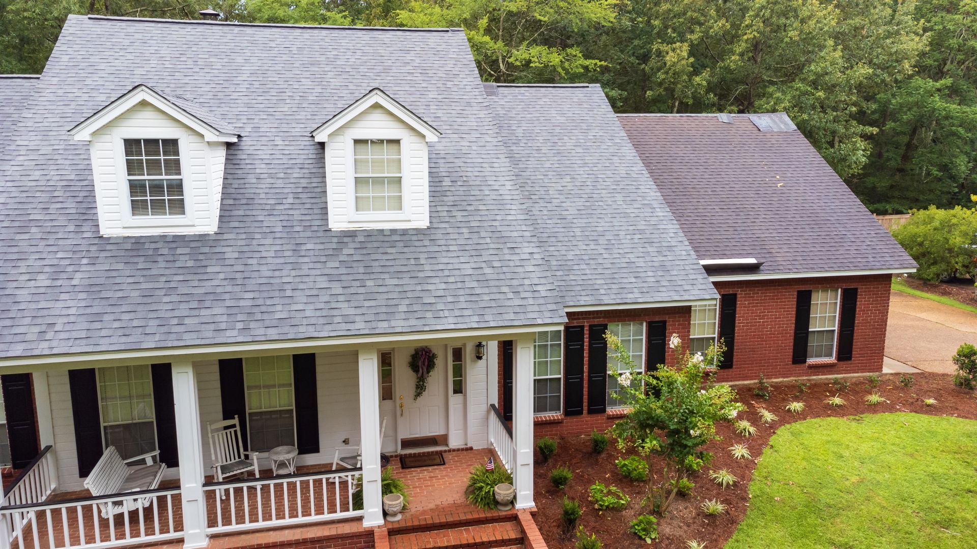 Brick house with gray roof, white porch, and manicured lawn surrounded by trees