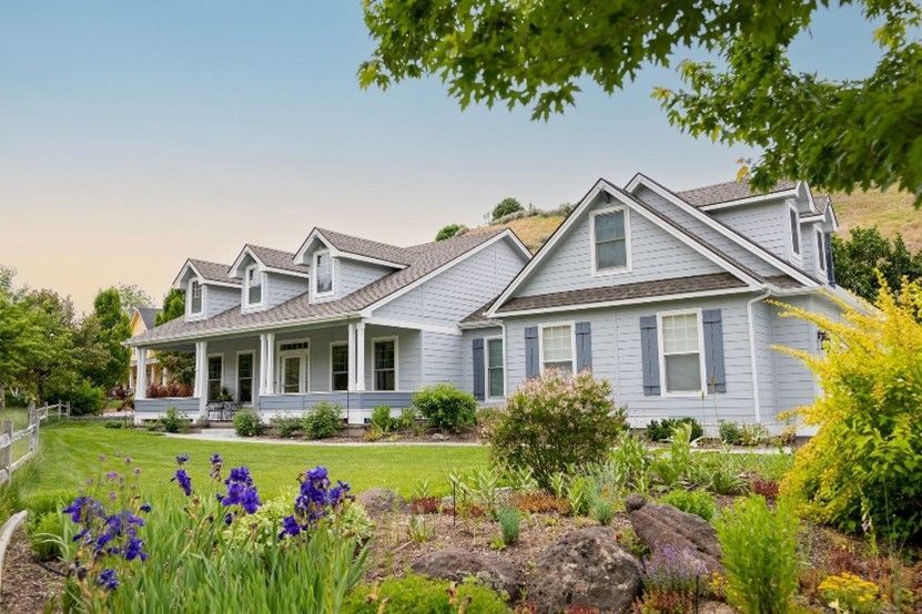 Gray farmhouse with white trim, porch, and purple flowers in a lush garden under green trees