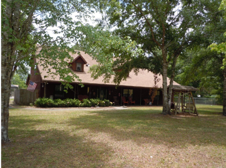 Rural house with a brown roof and front porch, surrounded by large trees and a grassy yard