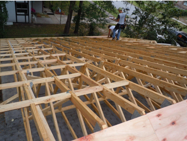Wooden roof framing under construction, with two workers in the background on a hillside site.