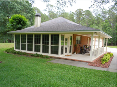 Small white screened porch with gray roof and chimney, surrounded by green lawn and trees.