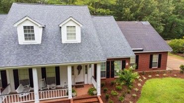 Gray-roofed brick house with white porch columns and dormer windows, surrounded by green lawn and trees