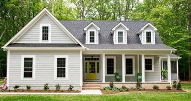 White two-story house with gray roof, dormer windows, and yellow front door, surrounded by green trees and lawn