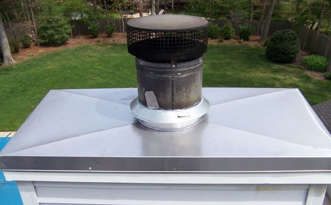Metal roof vent chimney on a silver rooftop with a black cap, set against a grassy backyard.