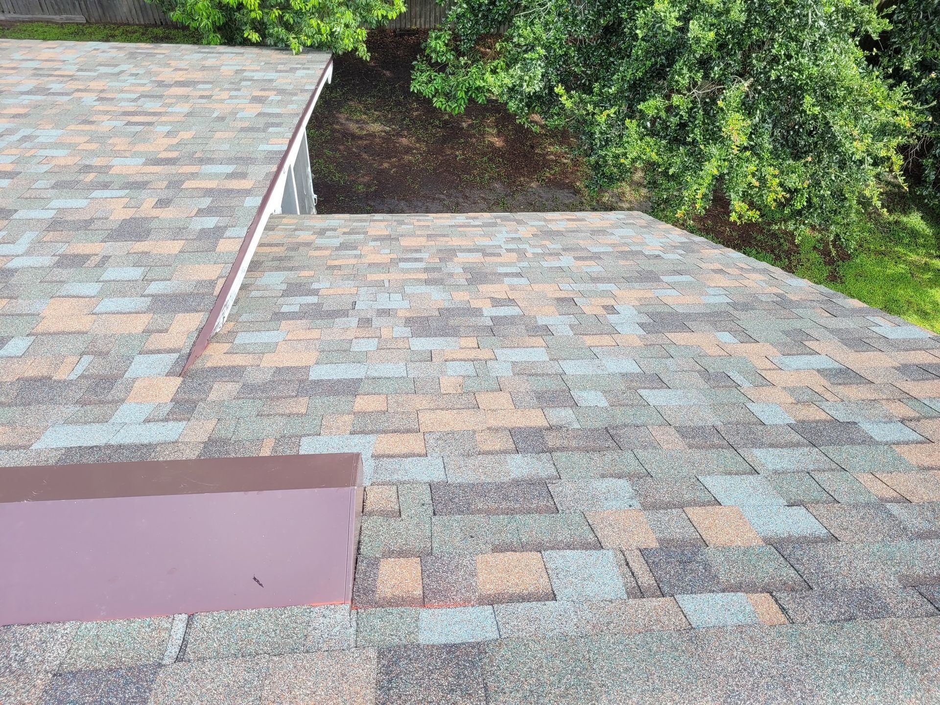 Overlapping shingled roof sections with a small vent and trees in the background
