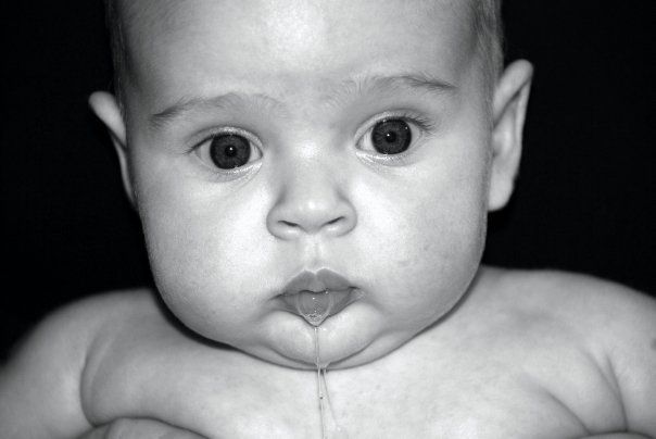 Black-and-white close-up of a baby with a drool string, looking straight at the camera.