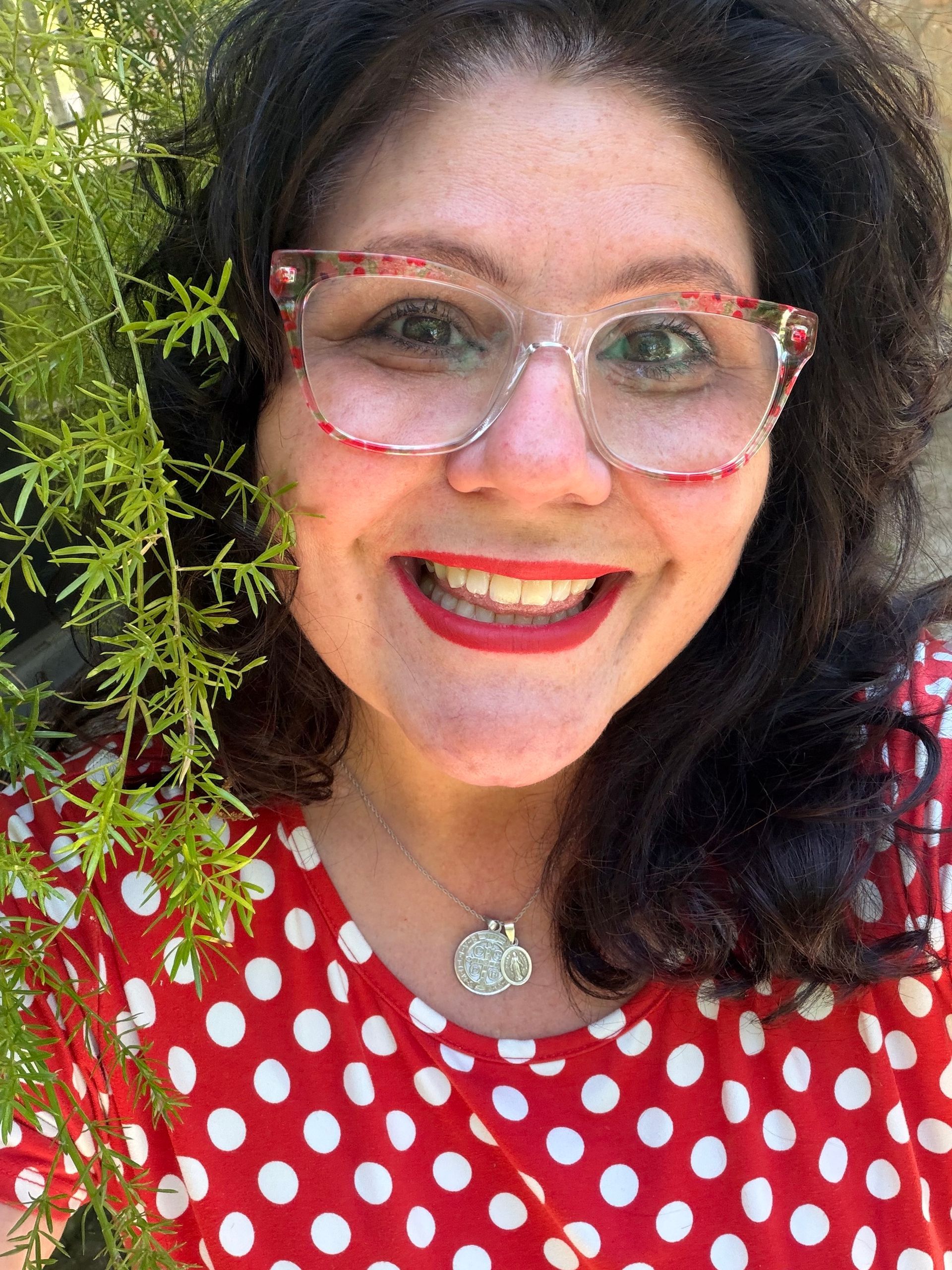 Smiling person with pink glasses and red polka-dot top, standing by green foliage