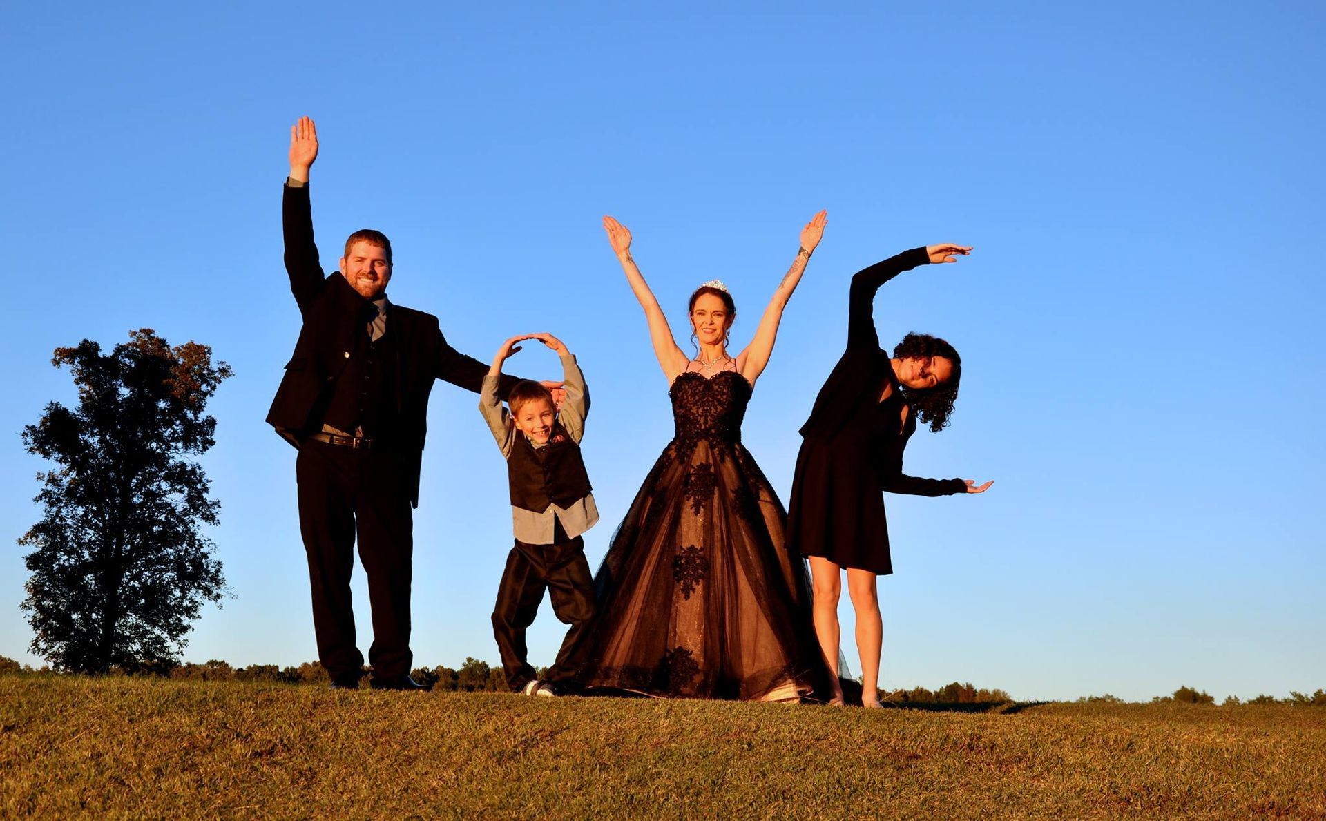 Four people posing on a grassy hill at sunset, arms raised against a blue sky.