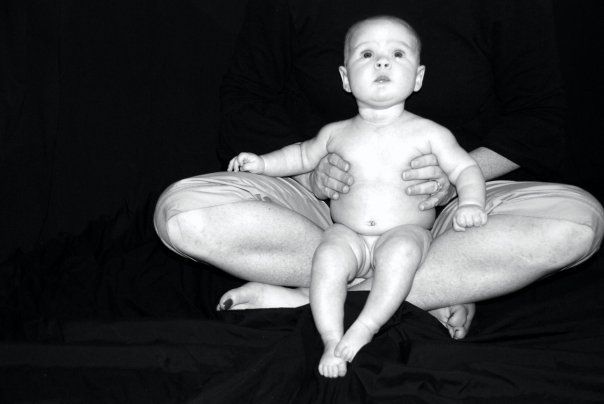 Black-and-white photo of a baby seated on an adult's crossed legs against a dark background.