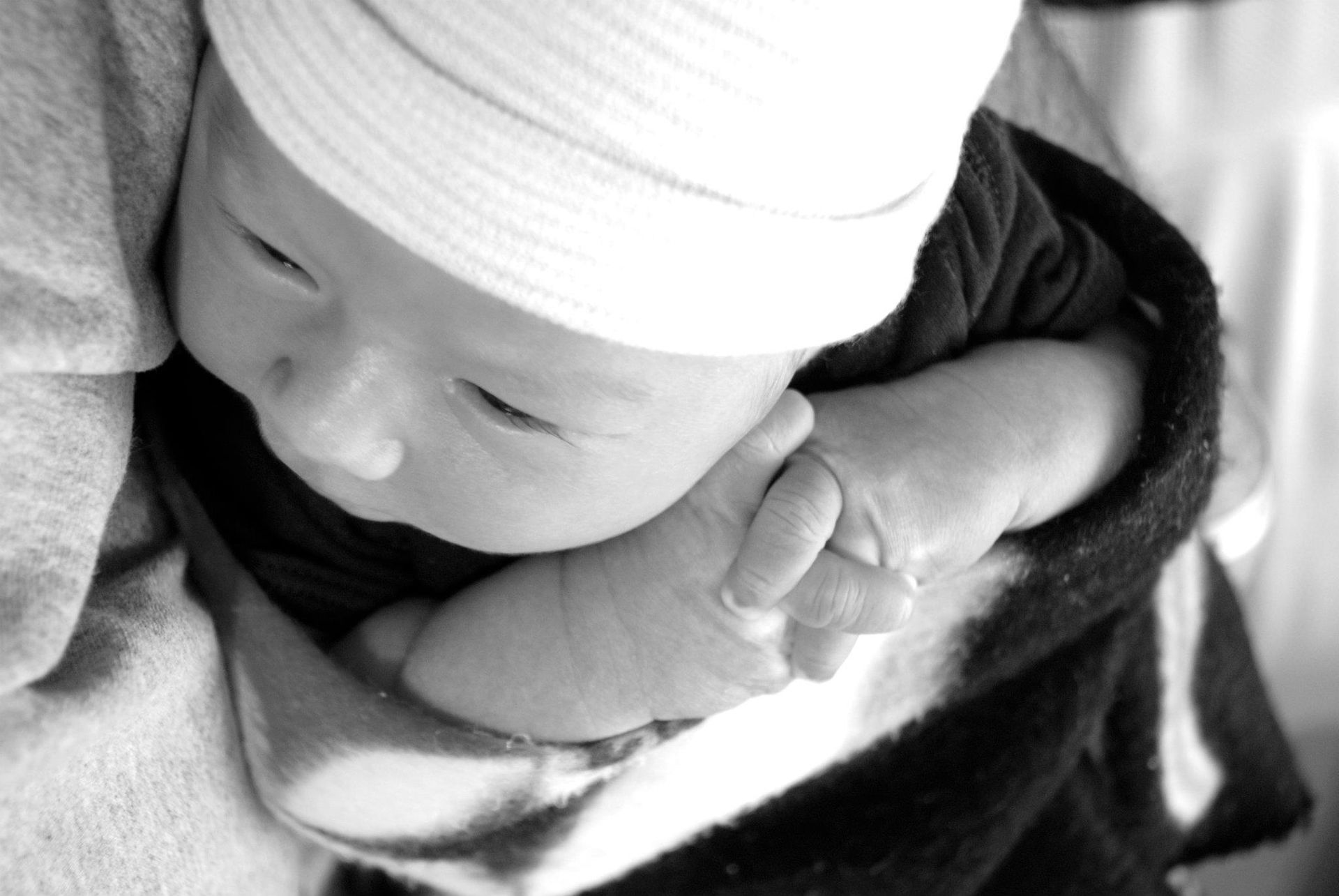 Sleeping baby wrapped in a dark blanket, wearing a white hat and resting on a patterned blanket.