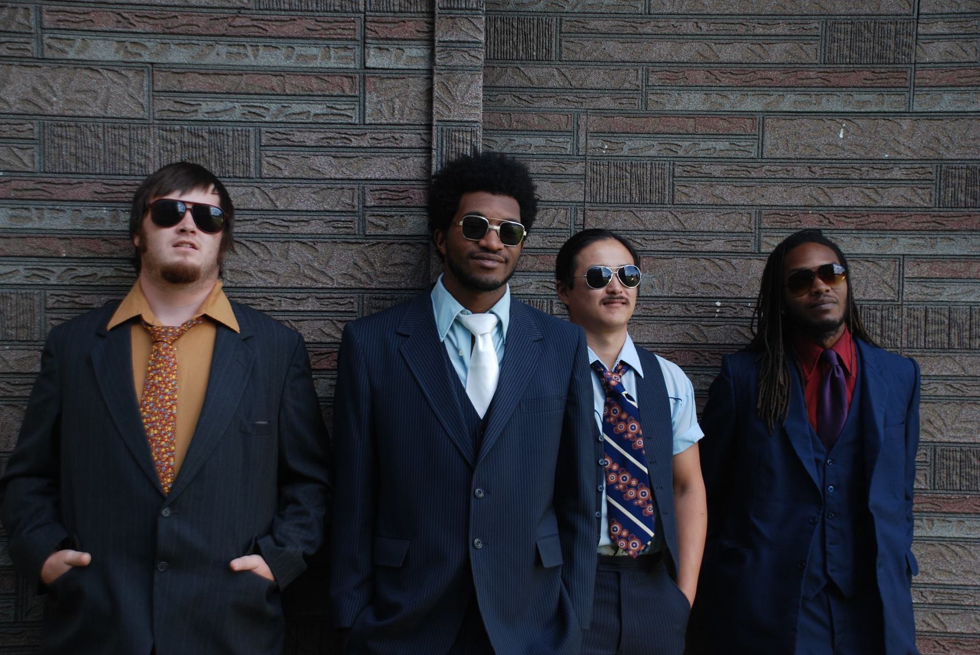 Four people in suits and sunglasses standing against a brick wall.