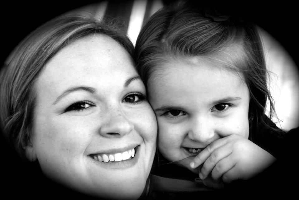 Smiling woman and child cheek-to-cheek in black and white, the child partly covering her mouth with a hand