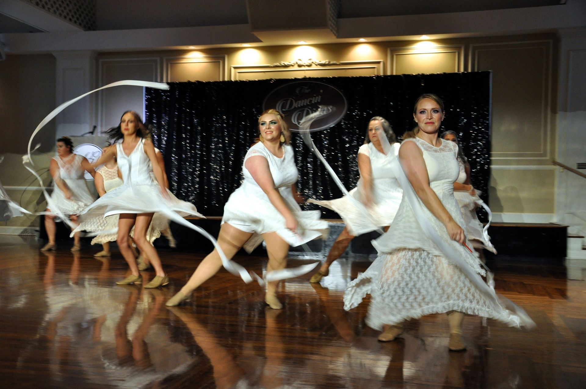 Dancers in white dresses perform a synchronized routine on a polished stage.
