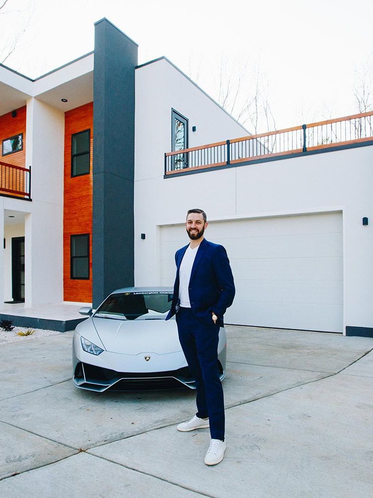 Man in blue suit stands in front of silver sports car and modern white house.
