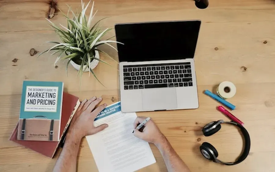 A person is writing on a piece of paper in front of a laptop computer.