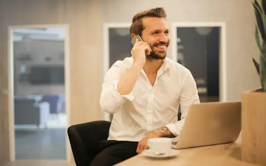 A man is sitting at a table with a laptop and talking on a cell phone.