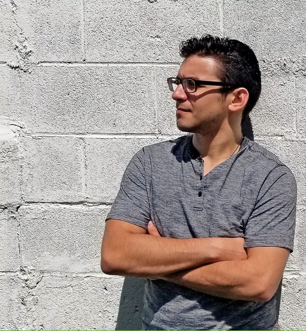 A man wearing glasses is standing in front of a brick wall with his arms crossed.