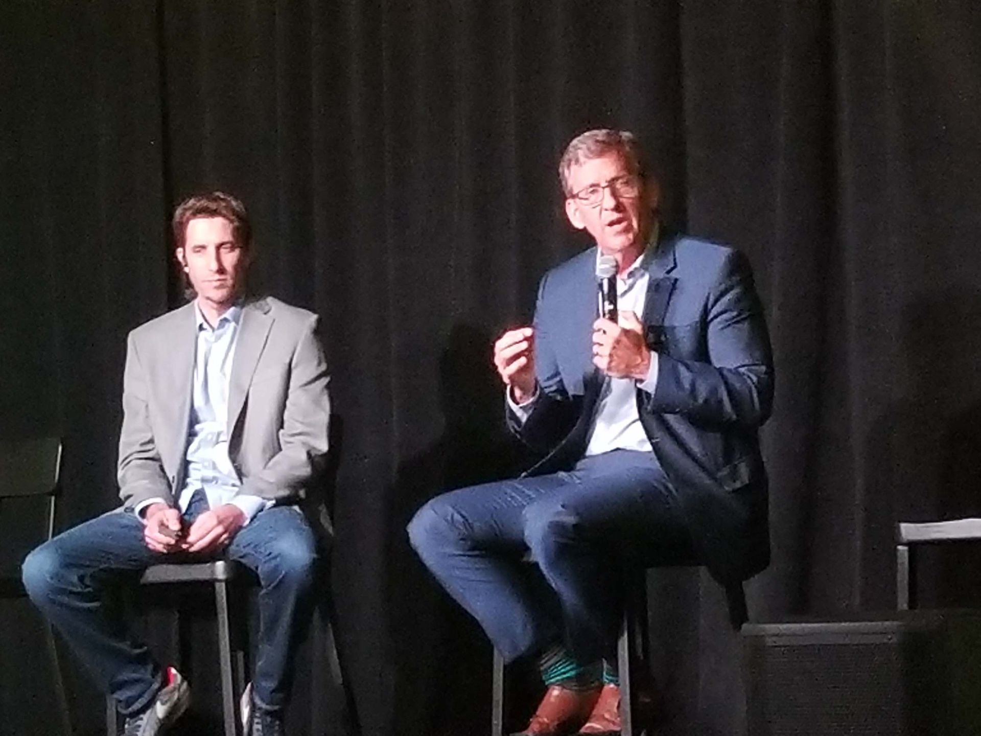 Two men are sitting on stools on a stage and one is holding a microphone