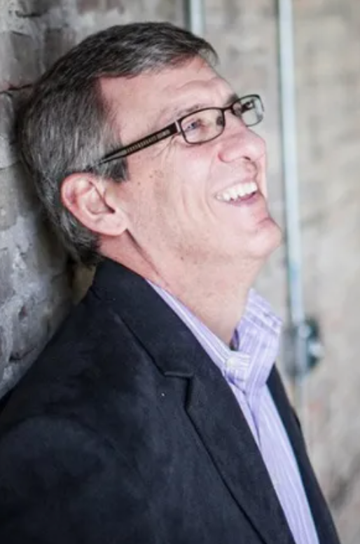 A man wearing glasses and a suit is leaning against a brick wall and smiling.