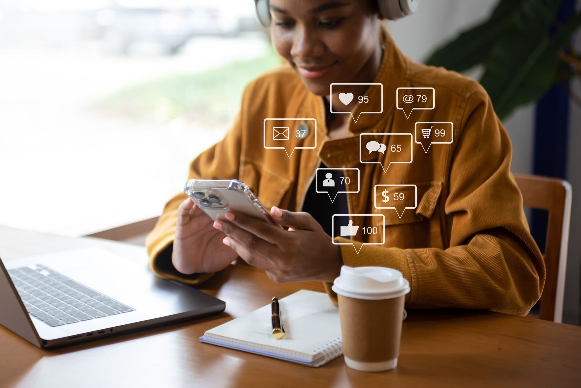 Person using a smartphone at a desk with a laptop, coffee, and social media icons hovering above.