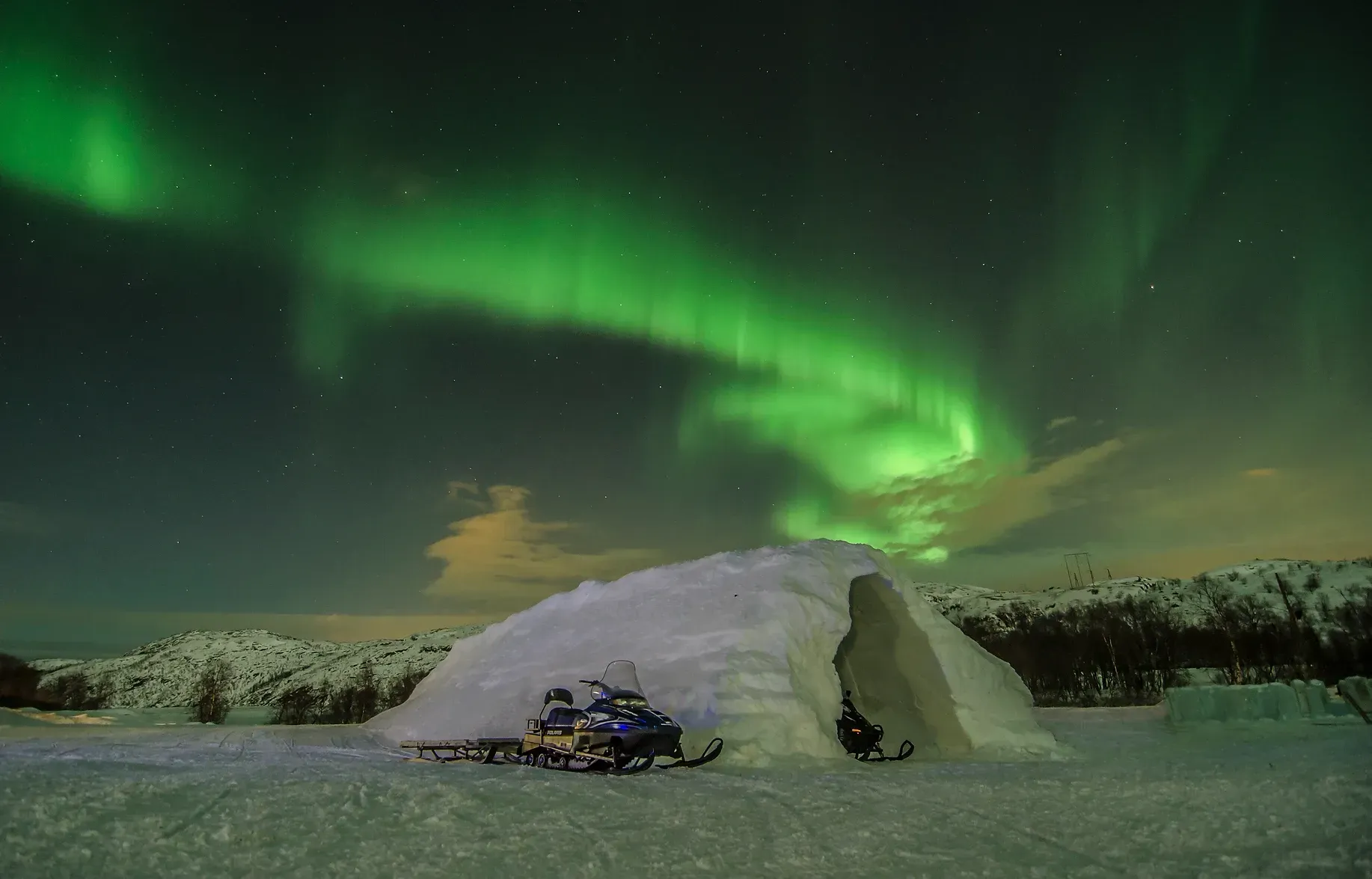 A snowmobile is parked in front of an igloo under the aurora borealis in Norway.