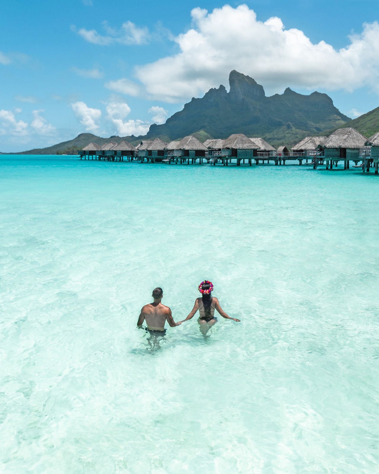 A man and a woman are holding hands in the ocean of Bora Bora with Four seasons overwater bungalows in the background. 