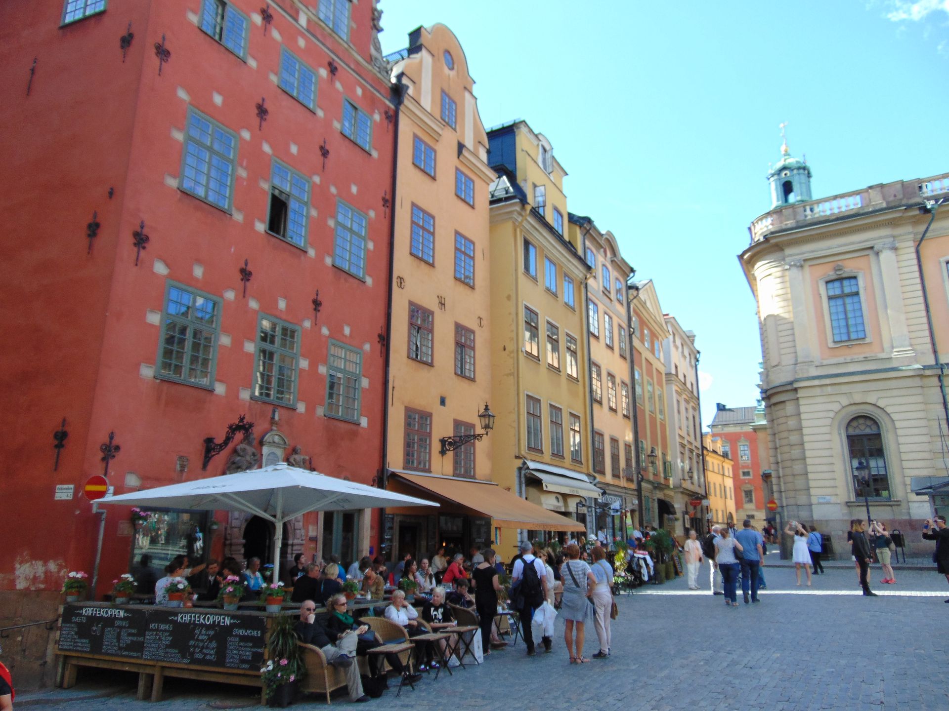 Colorful buildings line a cobblestone street with people at outdoor cafes in Stockholm, Sweden.