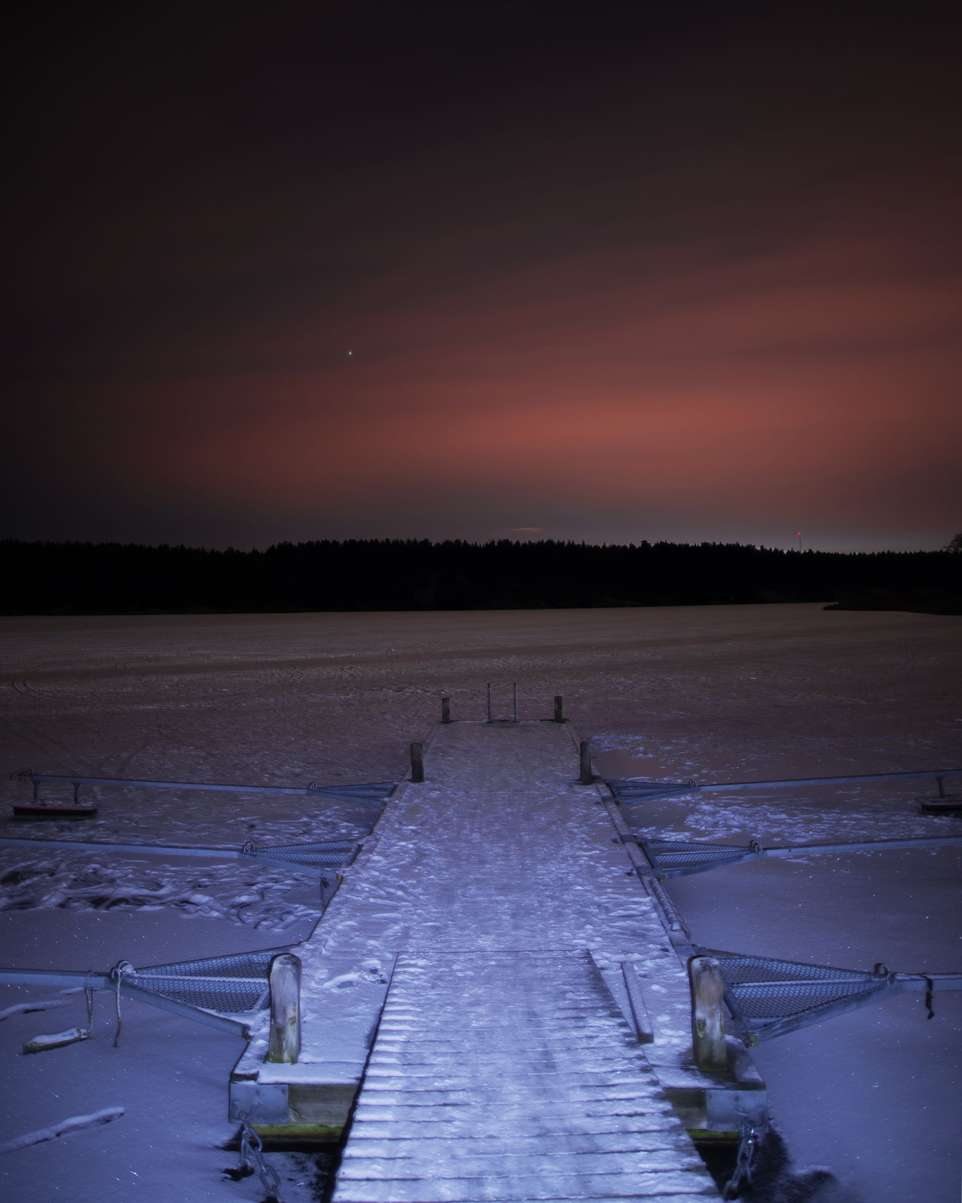 Wooden dock extends onto a frozen lake under a dusky, pink-tinged sky.