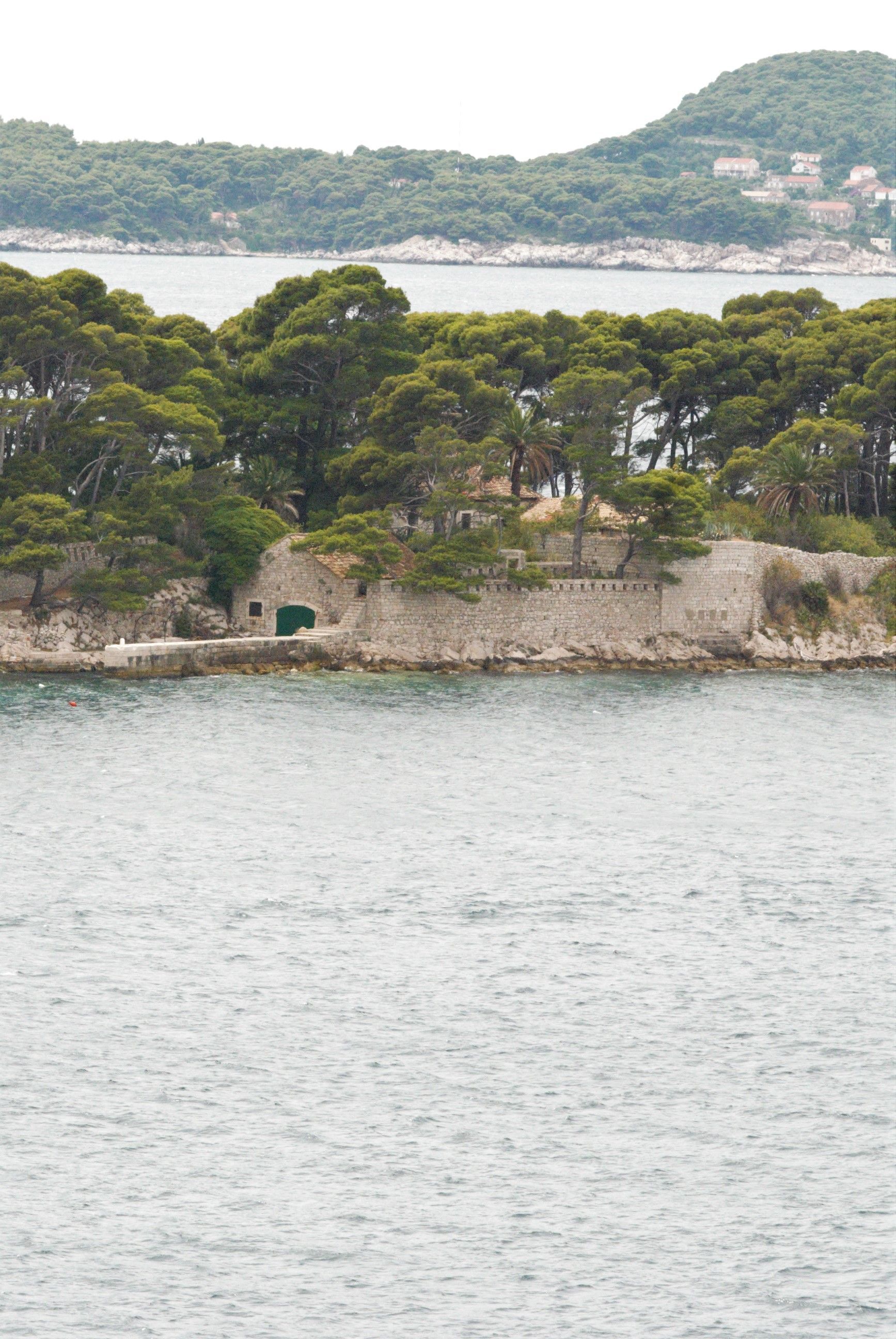 Coastal view: Stone wall with trees, green door, water, island with houses in the background.