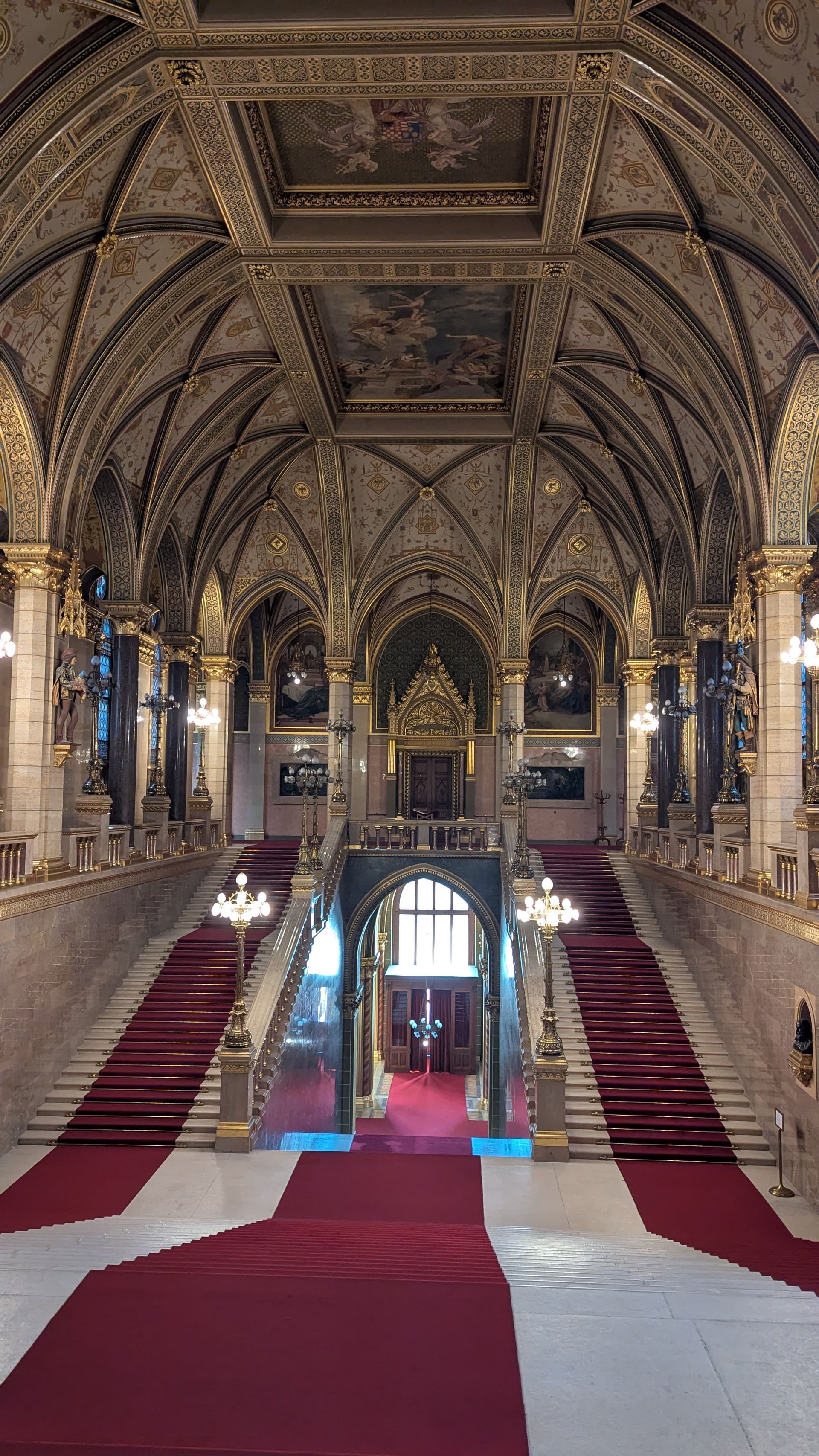 Ornate hall with grand staircase, red carpet, gold accents, and arched ceiling.